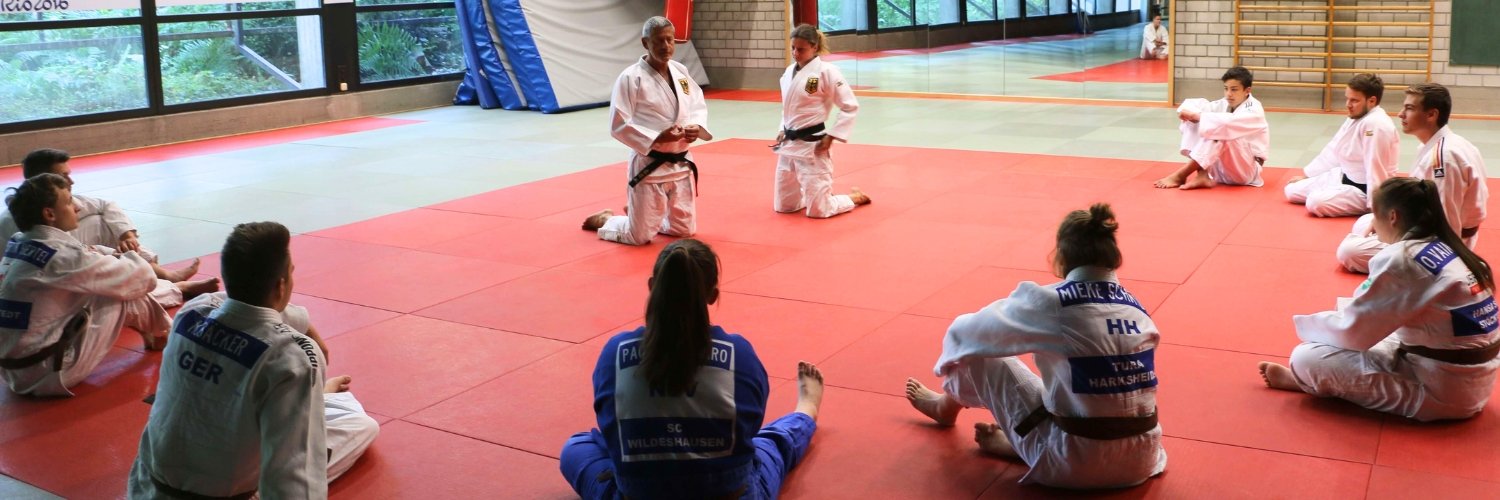 Judoka sitzen im Kreis auf dem Boden. Ein Judoka erklaert etwas.