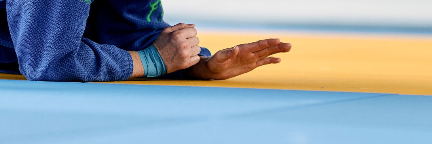 Detailaufnahme von den Haenden eines Judoka, der sich auf dem Boden abstuetzt. Um das rechte Handgelenk hat er blaues Tape gewickelt.