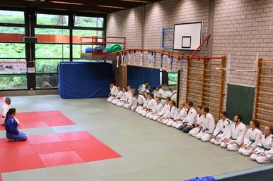 Viele Judoka sitzen in einer Reihe an der Wand einer Sporthalle beim Sportabzeichen. Vor ihnen sitzen zwei Judotrainer.